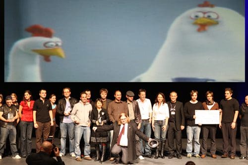 A diverse group of people, including men and women, standing on stage in front of a large screen displaying an animated chicken character. They are celebrating an achievement, holding awards and prizes.