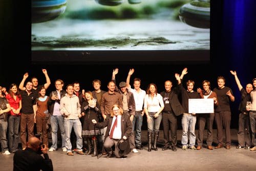 A diverse group of people, including men and women, celebrating on stage with arms raised, smiling and posing for a group photo. A large screen in the background displays a scenic image.