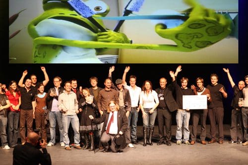 A large group of people on stage celebrating an event, with several raising their hands in excitement. A cartoon character is projected on a screen behind them, featuring a green creature holding a toothbrush.