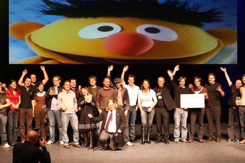 A diverse group of people smiling and celebrating on stage, with some raising their arms in joy. In the background, a large image of a character with an orange nose and large eyes is displayed on a screen.