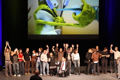 A diverse group of people gathered on stage, smiling and raising their arms in celebration. In the background, a large screen displays a vibrant image of a green cartoon character with blue toothbrushes.