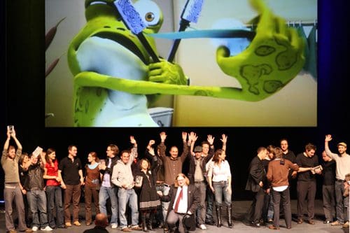 A group of diverse individuals on stage celebrating with raised hands, while a large animated character appears on a big screen in the background, holding a toothbrush. The atmosphere is festive and lively.