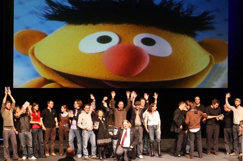 A large group of people gathered on stage, smiling and raising their hands in celebration. Behind them is a giant screen displaying a colorful image of a puppet with a big nose and spiky hair, creating a cheerful atmosphere.