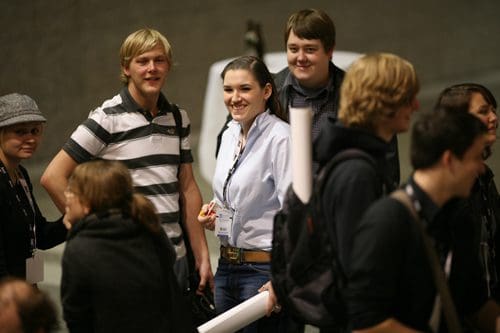 A group of young adults interacting in a casual setting, some smiling and holding posters. The scene captures a lively atmosphere, with individuals dressed in a mix of casual attire, suggesting a social or collaborative event.