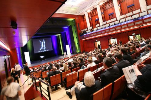 A large auditorium filled with an audience seated in rows of orange and brown chairs, facing a stage displaying a presentation. Colorful lights illuminate the backdrop, creating a vibrant atmosphere.