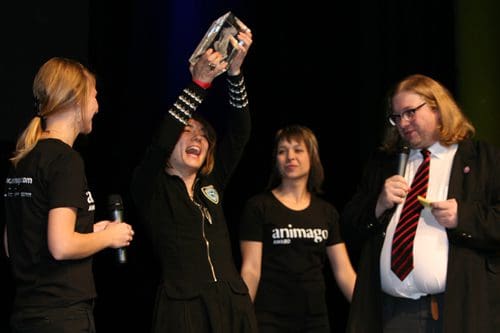 A woman in a black outfit joyfully raises a trophy above her head on stage, surrounded by three individuals in matching black t-shirts. The audience is engaged as they celebrate an achievement during an award ceremony.