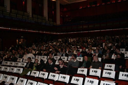 A large audience seated in a spacious auditorium with rows of empty seats. People are engaged in conversation, scattered throughout the venue, while light illuminates the stage area at the front.