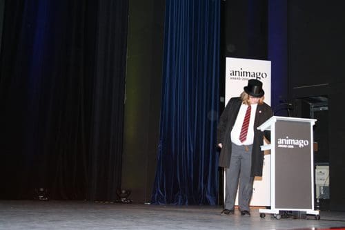 A man in formal attire, wearing a top hat, stands beside a podium with the logo of "animago" on it. He appears to be looking down, possibly adjusting his outfit. The background features dark curtains, suggesting a stage setting.