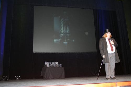 A man in a black top hat and formal attire stands on stage beside a display of awards. A projection screen behind him shows a blurred image. The stage is dimly lit, with a focus on the figure and awards.