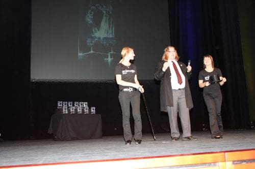 Three presenters on stage at an event, one in a black coat and tie holding a microphone, the others in black shirts. A table with trophies is in the background, under dim stage lighting.