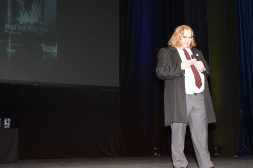 A man with long hair wearing a black coat and tie stands on a stage, adjusting his shirt. The backdrop features a dark display, and the stage floor is illuminated.