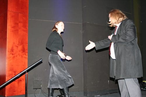 A woman wearing a black top and long gray skirt approaches a man in a black coat and white shirt, who extends his hand in greeting on a stage with a dark backdrop.