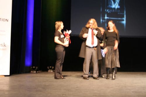 A stage scene featuring two women and a man with long hair, dressed in formal attire. The man holds an award while the women are receiving bouquets and engaging with him, set against a backdrop of soft stage lighting.