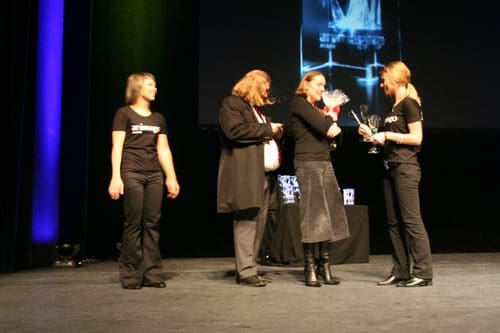 A group of individuals on stage at an awards ceremony. Two women in black outfits are receiving trophies from a man in a coat. A third woman stands nearby, watching the presentation. The backdrop features a lit display.