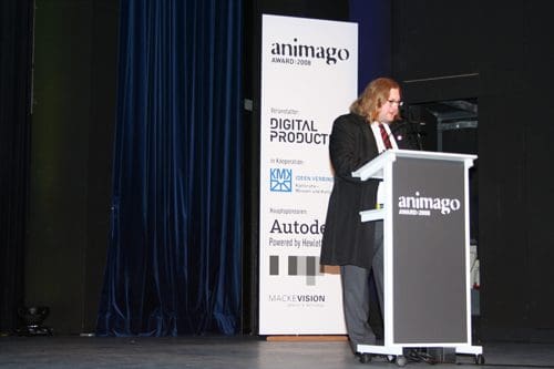 A speaker at a podium during the Animago Award ceremony, wearing a formal suit and glasses, with banners in the background promoting digital products and sponsors such as Autodesk and Mackevision.