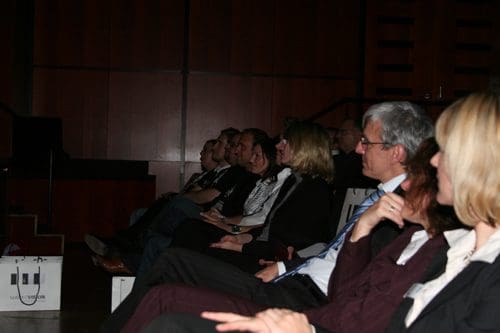 An audience seated in a dimly lit auditorium, focusing on a performance. They display a mix of attentive expressions, dressed in formal and business attire, set against a backdrop of wooden paneling.