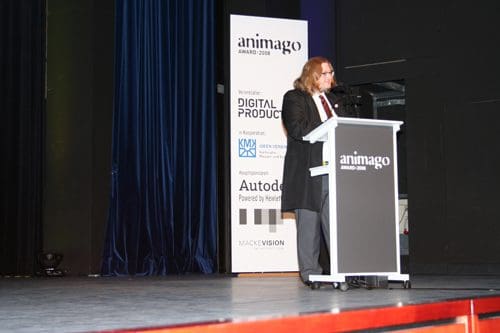 A person with long hair in a black suit standing at a podium during the animago awards ceremony, with a dark blue curtain in the background and event signage visible nearby.