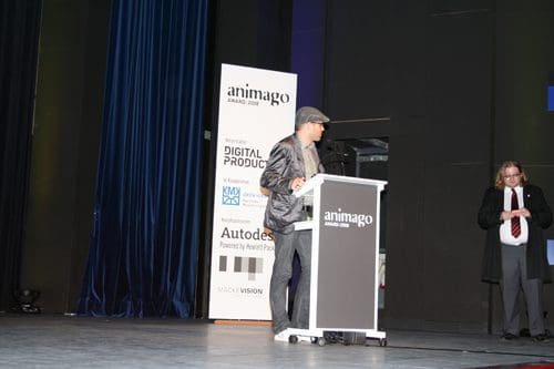A speaker in a dark suit and hat stands at a podium during the Animago Award ceremony, with a blue curtain in the background. Another individual in a suit observes to the right, while banners list sponsors and event details.