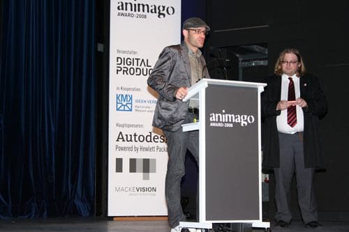 A man in a gray suit and cap stands at a podium delivering a speech during the animago Award 2008 event, while another man in formal attire observes nearby. A backdrop features event sponsors.