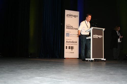 A man in a white shirt stands at a podium during a presentation at an event, with a backdrop displaying sponsors including Autodesk and animago, and a dark curtain behind him.