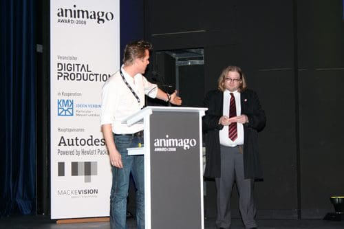 Two presenters on stage at the animago Award 2008 ceremony. One man, dressed casually in a white shirt and jeans, gestures as he speaks, while the other man, in a suit with a red tie, listens attentively. A banner featuring event sponsorship is visible in the background.