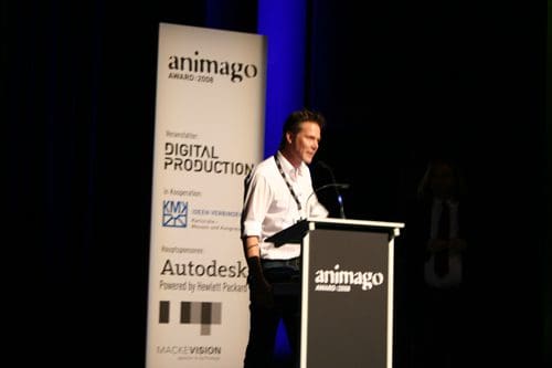 A man standing at a podium, speaking at the animago awards event. Behind him, a backdrop features logos from sponsors like Autodesk and Digital Production. The setting is a stage with dim lighting, focusing on the speaker.