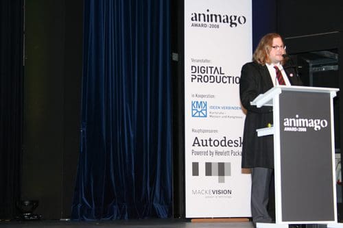 A speaker in a black suit presenting at a podium during the Animago Award 2008 event, with a backdrop featuring sponsors like Autodesk and Digital Production. Blue curtains drape in the background.