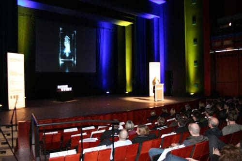 An auditorium filled with an audience seated in rows, watching a speaker at a podium. A large screen behind displays a black-and-white image. The stage is illuminated with blue and green lights.