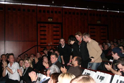 An audience applauding as a group of people walks towards the stage in a theater. The setting features wooden walls and rows of seated guests, showcasing a moment of recognition or celebration.