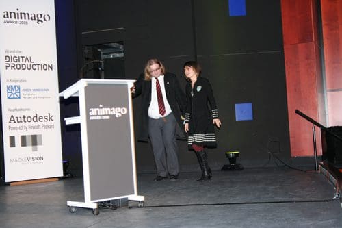 A man in a suit with long hair and a woman in a black dress with striped patterns walk together on stage at the animago award ceremony, with a podium displaying the event name. The venue is dimly lit, highlighting the stage area.