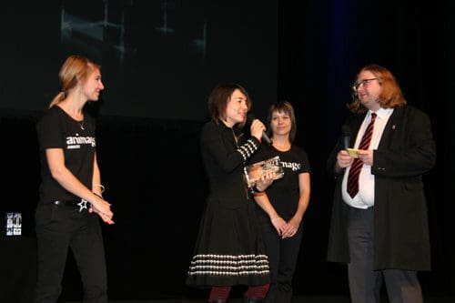 A presenter holds an award on stage while speaking to an audience. Two women in black t-shirts with "animago" printed stand beside her. A man in formal attire observes as they celebrate the achievement during an event.