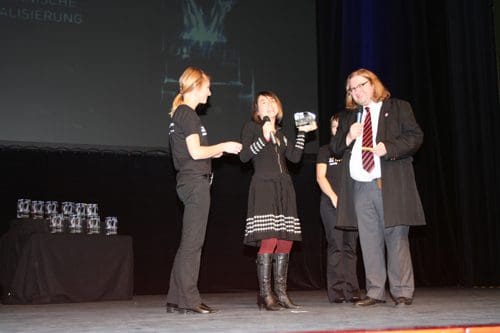 A woman in a black dress stands on stage with a microphone, engaging with an audience member. Two individuals in black shirts stand nearby, while a man in a suit with glasses observes. Trophies are displayed on a table behind them.