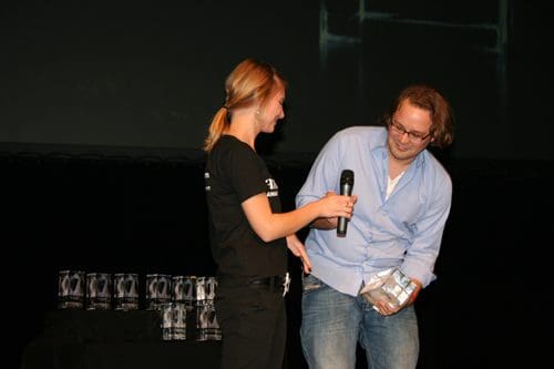 A woman in a black shirt holding a microphone presents an award to a man in a light blue shirt at a stage event. In the background, several trophies are visible on a table.
