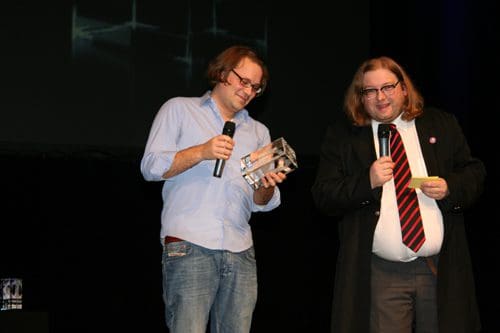 Two men on stage at an event; one man is holding a trophy and microphone, while the other is speaking into a microphone. The background features a projection with abstract patterns.
