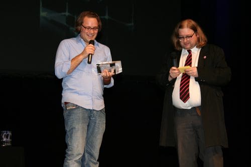 Two men on stage during an event. One man holds a microphone and a clear award, smiling, while the other, dressed in a long black coat and tie, reads from a yellow note. A dark backdrop with a projection is visible.