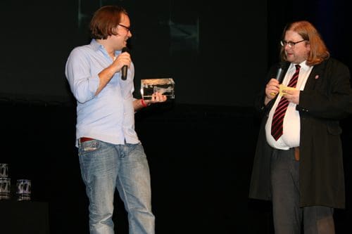 Two men are standing on stage in front of a dark backdrop. One man, wearing a light blue shirt and jeans, is holding a trophy and microphone, smiling. The other man, dressed in formal attire with a black coat and red tie, is also holding a microphone and laughing.