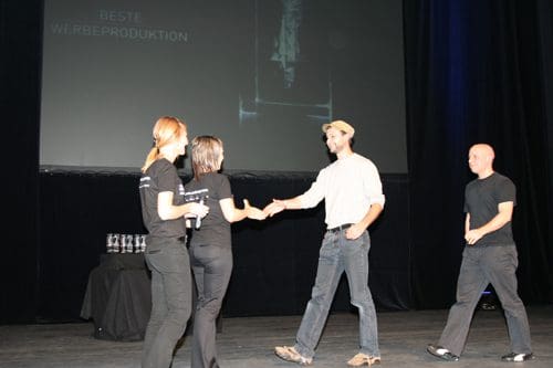 A man in a white shirt shakes hands with a woman as part of a presentation on stage, while two other individuals observe. The backdrop displays text in German and there are awards on a table nearby.