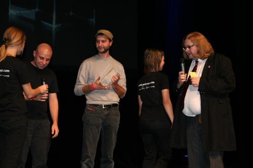 A group of individuals on stage at an event, with two people conversing while holding microphones. One person is in a black coat, another is holding a glass trophy, and others are wearing black t-shirts, engaged in casual conversation.
