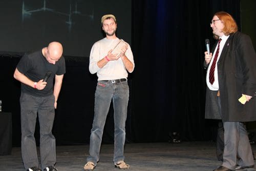 Two men on stage during an event, one is bowing while holding a small trophy, and another man in a suit is speaking into a microphone. The backdrop is dark with an illuminated screen.