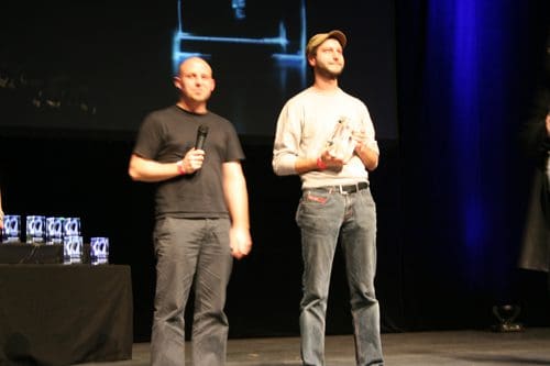 Two men standing on a stage during an awards ceremony. One man, holding a microphone, is wearing a black shirt and gray pants, while the other man, wearing a light-colored shirt and cap, is holding an award. Behind them, a table is set with trophies.
