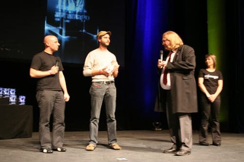 Two men stand on stage during an award ceremony. One man holds a trophy, while the other, dressed in a black coat, speaks. A woman in a black shirt watches from the side. The background features colorful stage lighting.