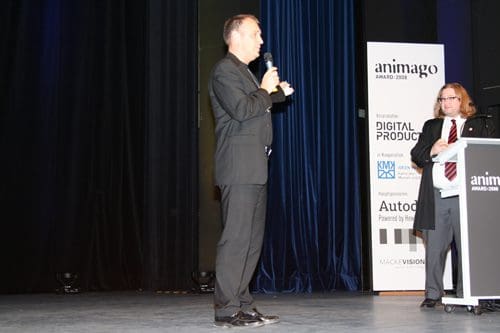 A speaker in formal attire holding a microphone on stage, addressing an audience at an awards ceremony. Another individual in a suit stands beside a podium with branding. The stage backdrop features dark curtains and event signage.