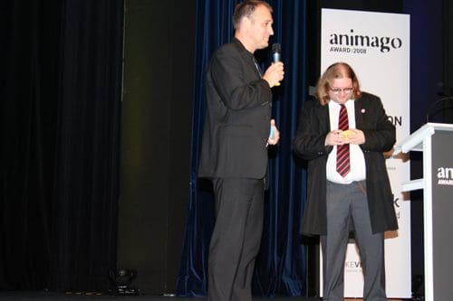 Two men on stage at an award ceremony. One man is speaking into a microphone, wearing a black suit, while the other, dressed in a suit with a red tie, is holding a device and appears to be listening.