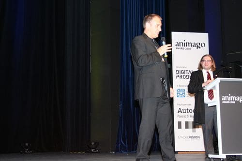 A man in a black suit speaking into a microphone on stage at an award ceremony. Another man in a black suit and glasses stands beside him, with a backdrop featuring logos of sponsors and the event name, animago Award 2008.
