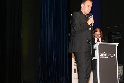 A man in formal attire stands at a podium with a microphone, speaking at an awards event. Another individual sits at the podium, partially obscured. The backdrop features dark blue curtains and an event sign for animago 2008.