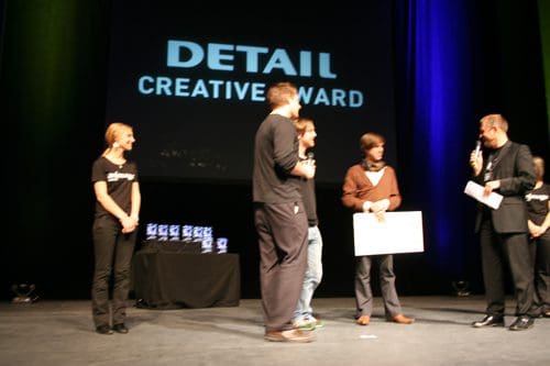 A stage setting for the DETAIL Creative Award ceremony, featuring a speaker holding a microphone and presenting an award to a group of smiling recipients. In the background, award trophies are displayed on a table, illuminated by colored stage lights.
