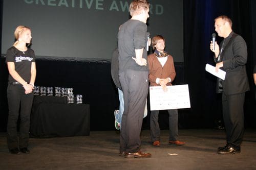 A stage scene during an awards ceremony, featuring a presenter speaking into a microphone, a recipient holding a large check, and a woman standing to the side. Awards are displayed on a table in the background.