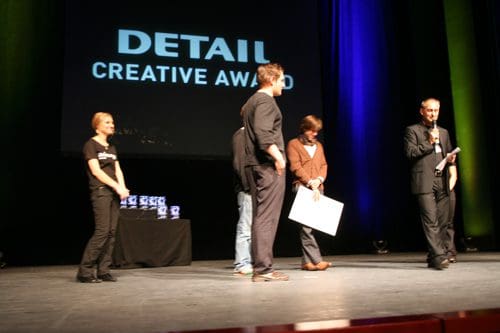 A group of people on stage at an awards ceremony. One man is speaking into a microphone while holding a paper, flanked by three others. Behind them is a screen displaying 'DETAIL CREATIVE AWARD' and trophies are visible on a table.