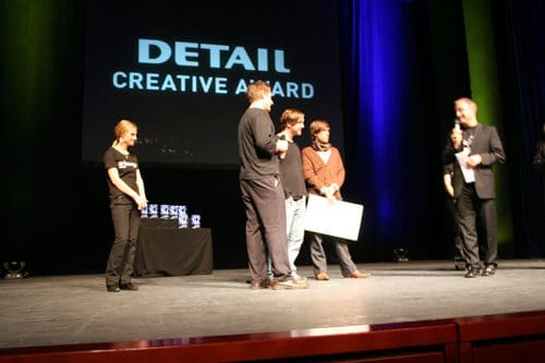 A group of four individuals on stage at an award ceremony, with one person presenting a large check. A backdrop displays the text "DETAIL CREATIVE AWARD". A table with trophies is set off to the side.