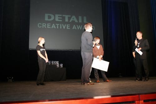 A stage during an award ceremony featuring four people. One person hands a large check to another while a woman stands on the side. Trophies are displayed on a table in the background with a large screen showing 'DETAIL CREATIVE AWARD'.